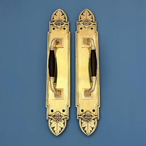 May include: Two brass door handles with black grips. Each handle has a rectangular backplate with leaf and scroll details. The handles have a curved design and are mounted on a blue background.