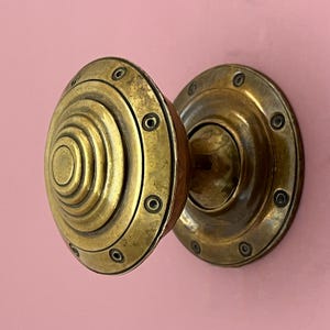 May include: A close-up shot of a brass door knob with a spiral design against a pink backdrop. The knob features a circular shape with concentric rings and is fixed to a round backplate. Small screws secure the knob in place.