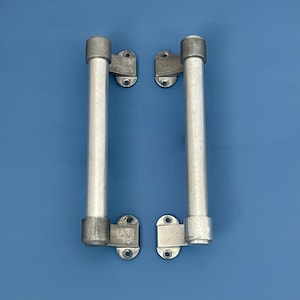 May include: Two silver-colored metal cabinet pulls with mounting plates. Each pull has a cylindrical handle and is attached to a rectangular plate with screw holes. The background is a solid blue.