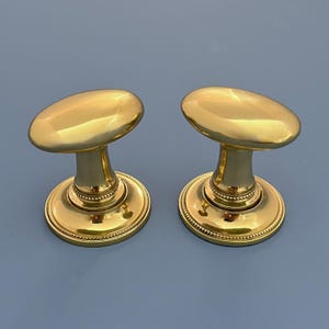 May include: A pair of gold-coloured door knobs. Each knob has an oval top and a round base with a beaded edge. The background is a light blue colour.