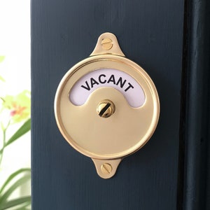 May include: A brass door sign with a round, rotating face that says "VACANT" in black lettering on a white background.
