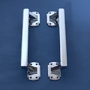 Peut inclure: Deux poignées de porte chromées polies. Chaque poignée a une barre rectangulaire et est montée sur une base rectangulaire décorative avec des coins arrondis. Les poignées sont posées sur un fond bleu foncé.