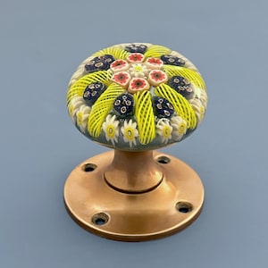 Puede incluir: Un pomo de puerta decorativo de cristal con diseño floral. El pomo presenta una cúpula de cristal transparente con motivos florales amarillos, rojos, blancos y azules. Está montado sobre una base metálica redonda de color bronce con cuatro orificios para tornillos.