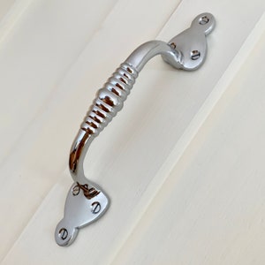 Può includere: Una maniglia per porta in cromo lucido con un design a coste. La maniglia ha una forma curva ed è fissata a una superficie bianca.