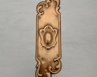 May include: A gold-toned metal door plate with a decorative, ornate design. The plate has a raised, oval-shaped knob in the centre.
