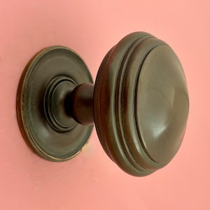 Könnte beinhalten: Ein bronzefarbener Türknauf mit einem runden, geriffelten Griff und einer kreisförmigen Rückplatte. Der Knauf ist auf einer rosa Oberfläche montiert, die sein antikes Design und seine metallische Oberfläche hervorhebt. Dieses Hardware-Teil verleiht einen Vintage-Touch.