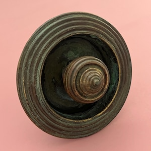 May include: A close-up of a bronze-colored, antique-style object with a circular design. The object has a central, spiraled knob and a surrounding, ridged rim. The piece has a weathered appearance, suggesting age and use.