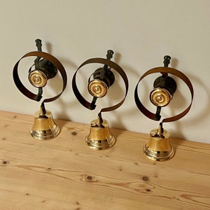 May include: Three antique brass bells with black metal rings. The bells are hanging from the rings and have a gold finish.