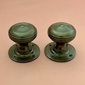 May include: Two matching dark bronze door knobs with a circular, ribbed pattern on the top surface. Each knob features a smooth, rounded handle attached to a circular base plate with four pre-drilled screw holes for mounting to a door surface.
