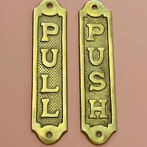 May include: Two rectangular brass door plates with rounded ends and mounting holes at the top and bottom. One plate features the word PULL in raised letters, and the other features the word PUSH in raised letters, both set against a textured background.