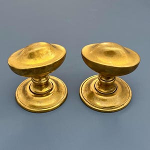 May include: Two antique brass door knobs. Each knob has a rounded, disc-shaped top and a cylindrical base. The brass has a warm, golden hue, and the knobs are set against a muted blue-gray background. These knobs are likely for interior doors.