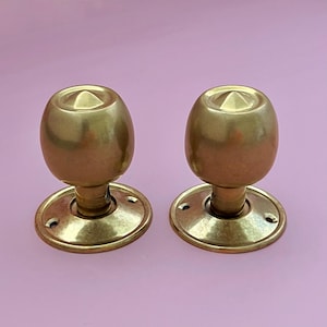Könnte beinhalten: Zwei passende Türknöpfe aus Messing mit polierter Metalloberfläche. Jeder Knauf hat eine abgerundete, bauchige Form mit einem kleinen, spitzen kegelförmigen Detail in der Mitte der Oberseite. Sie sind an runden Montageplatten mit Schraublöchern befestigt.