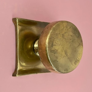 Könnte beinhalten: Ein Vintage-Türknauf aus Messing mit rundem Griff und quadratischer Rückplatte. Der Türknauf hat ein abgenutztes, antikes Aussehen mit leicht angelaufenem Finish. Die Hardware ist auf einer rosa Oberfläche montiert.