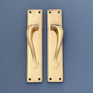 Puede incluir: Dos manijas de puerta de latón pulido con placa trasera rectangular y diseño de manija curva. Cada manija tiene un detalle decorativo en forma de corazón en la base. Fondo azul.