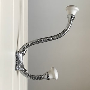May include: A chrome coat hook with a white ceramic knob on each end. The hook is curved and has a decorative design.