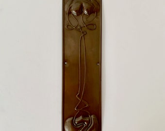 May include: A brown metal door plate featuring an ornate floral design. The plate is rectangular and decorated with a stylised flower with a long stem.