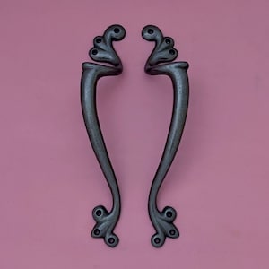 May include: A pair of black metal door handles with ornate, decorative details. The handles have a curved shape and a floral design.