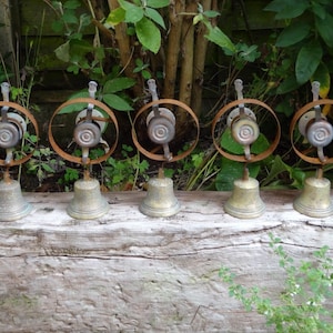 May include: Five antique brass bells with a circular metal design. The bells have a weathered appearance, with a rusty brown metal ring around the top. The bells are arranged in a row on a wooden surface, with greenery in the background.