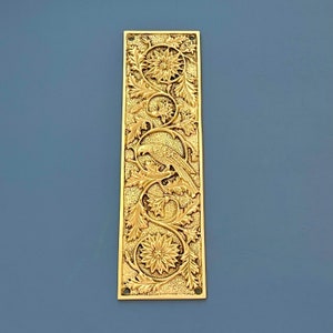 Op de afbeelding: Een rechthoekige messing deurplaat met een reliëf ontwerp. Het ontwerp toont een vogel die op een bladrijke tak zit, omringd door bloemen en wervelende patronen. De plaat heeft een warme goudkleur en een vintage uitstraling.