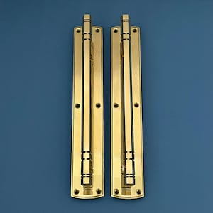 Puede incluir: Dos tiradores de puerta de latón con placa trasera rectangular y asa vertical. Los tiradores son de color dorado brillante y tienen un diseño clásico. Cada tirador tiene una serie de anillos decorativos y una parte superior cuadrada. El fondo es azul liso.