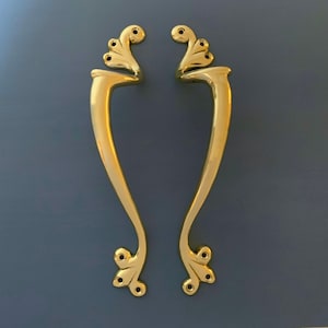 Può includere: Un paio di maniglie per porte dorate ornate con un design decorativo a spirale. Le maniglie hanno una finitura liscia e lucida.