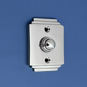 May include: A polished chrome doorbell button with a rounded, raised button. The button is surrounded by a decorative frame with a rectangular shape.