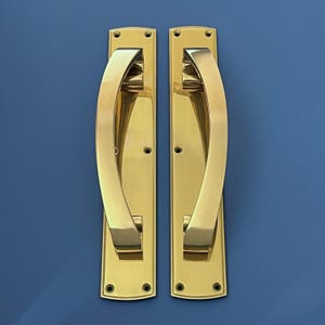Puede incluir: Dos manijas de puerta de latón pulido con un diseño curvo. Cada manija está montada en una placa rectangular con esquinas redondeadas y orificios para tornillos. El conjunto se encuentra sobre un fondo azul liso.