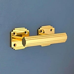 Può includere: Una maniglia per porta decorativa color oro, di forma rettangolare e con un design scanalato. La maniglia ha una base quadrata con angoli arrotondati e due fori per viti visibili.