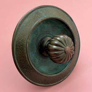 May include: A bronze-toned door knob with a decorative, ridged handle and a circular backplate. The backplate has a textured outer ring and a darker, aged patina. The knob is mounted on a pink wall.