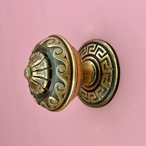 Könnte beinhalten: Ein runder Messing-Möbelknopf mit einer zentralen, geriffelten Kuppel, die von einem erhabenen Schnörkelmuster umgeben ist. Die kreisförmige Grundplatte ist mit einem geometrischen Mäander-Muster verziert und hat eine dunkle Patina.