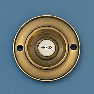 Könnte beinhalten: Ein runder, messingfarbener Türklingelknopf mit einem weißen, runden Knopf, auf dem "PRESS" in schwarzen Buchstaben steht.