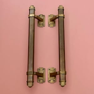 Könnte beinhalten: Zwei antike Türgriffe aus Messing mit rechteckigen Montageplatten und langen, achteckigen Griffen. Die Beschläge haben eine warme, gealterte Messingfarbe, wahrscheinlich im Art-Deco-Stil. Die Türgriffe sind an einer rosa Wand montiert.