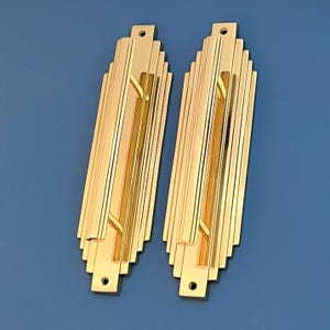 Puede incluir: Dos tiradores de puerta decorativos de color dorado con un diseño Art Deco. Cada tirador tiene una forma rectangular con una manija empotrada y un patrón geométrico escalonado.