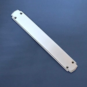 Puede incluir: Un tirador de puerta plateado con un borde decorativo festoneado. El tirador es largo y rectangular con dos agujeros para tornillos en cada extremo.