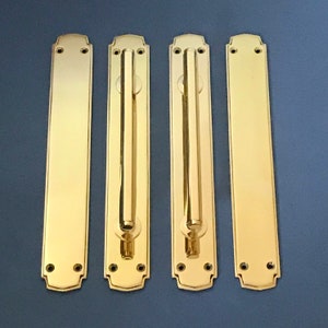 Pode incluir: Quatro puxadores de porta dourados com forma retangular e uma longa pega vertical. Os puxadores têm um design decorativo e ornamentado em torno das bordas.