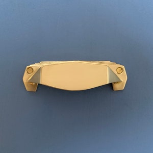 Può includere: Una maniglia per armadio dorata con un design geometrico. La maniglia ha una forma rettangolare con bordi arrotondati e un centro leggermente rialzato.