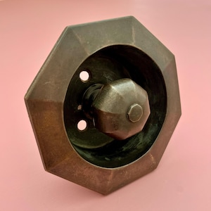 May include: A bronze-colored, octagonal door knob with a matching faceted knob. The knob is set within a recessed, octagonal plate. Two small holes are visible near the knob. The hardware has a vintage aesthetic.