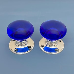 May include: Two blue glass door knobs with a polished chrome base. The knobs are a deep, vibrant blue and have a rounded, spherical shape. The base is circular with three screw holes. The door knobs are set against a light blue background.