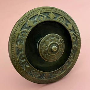 May include: Antique brass-toned cabinet knob with a circular design. The knob features an ornate, embossed pattern around the outer edge and a central, smaller knob with a floral design. The knob is likely made of metal and has a dark, possibly painted, center.