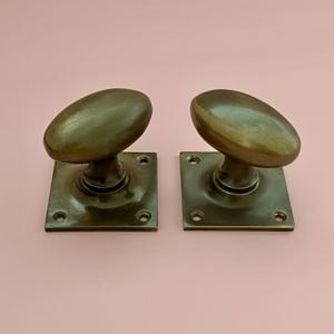 Peut inclure: Une paire de poignées de porte ovales avec une finition en bronze foncé. Chaque poignée est fixée sur une plaque carrée en métal dotée de quatre trous de vis pré-percés aux coins. Le métal présente une surface lisse et légèrement réfléchissante.