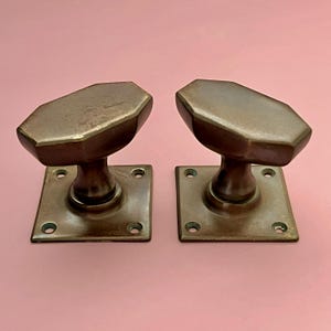 Pode incluir: Um par de maçanetas de porta correspondentes com um formato octogonal alongado e acabamento em bronze escuro. Cada maçaneta está fixada a uma placa de montagem quadrada com quatro orifícios para parafusos pré-perfurados nos cantos.