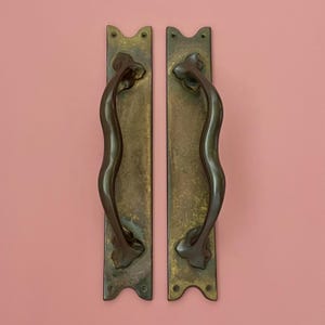 Puede incluir: Dos manijas de puerta antiguas de bronce. Cada manija tiene una placa trasera larga y rectangular con un diseño floral decorativo en la parte superior e inferior. La manija curva es de color bronce oscuro. El fondo es rosa liso.