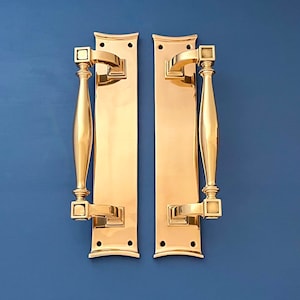 May include: A pair of polished brass door handles with a rectangular backplate and a curved top. The handles have a square design at the top and bottom.