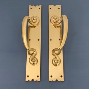 May include: A pair of polished brass door handles with ornate, curved designs. The handles are attached to rectangular plates with decorative edges.