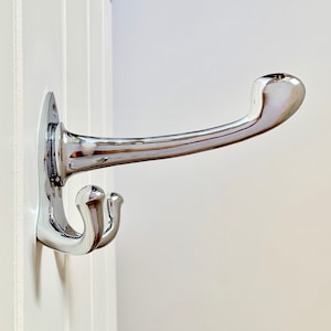 Puede incluir: Un gancho para abrigos cromado con un solo gancho y un brazo curvo. El gancho está montado en una pared blanca.