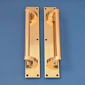 Puede incluir: Un par de manillas de puerta de latón pulido con un agarre cilíndrico largo y un diseño curvo en la parte superior e inferior. Las manillas están montadas en placas rectangulares con agujeros de tornillo visibles.