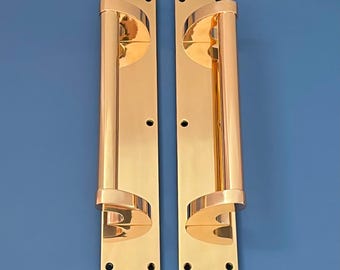 Könnte beinhalten: Zwei polierte Messing-Türgriffe mit einem langen, zylindrischen Griff und einem geschwungenen Design oben und unten. Die Griffe sind auf rechteckigen Platten mit sichtbaren Schraubenlöchern montiert.