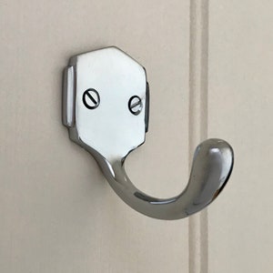 May include: A chrome-plated hook with a hexagonal base and a rounded hook. The hook is mounted on a white wall.