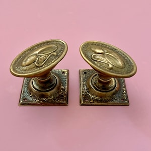 Op de afbeelding: Twee antieke messing deurklinken met een ovaal ontwerp en vierkante basis. De handgrepen hebben een gedetailleerd, getextureerd oppervlak en staan tegen een roze achtergrond. Het ovale ontwerp heeft een gestileerd bloemen- of organisch motief.