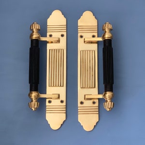 May include: A pair of gold door handles with black, ribbed, decorative pulls. The handles have a traditional design with a rectangular plate and a decorative top.